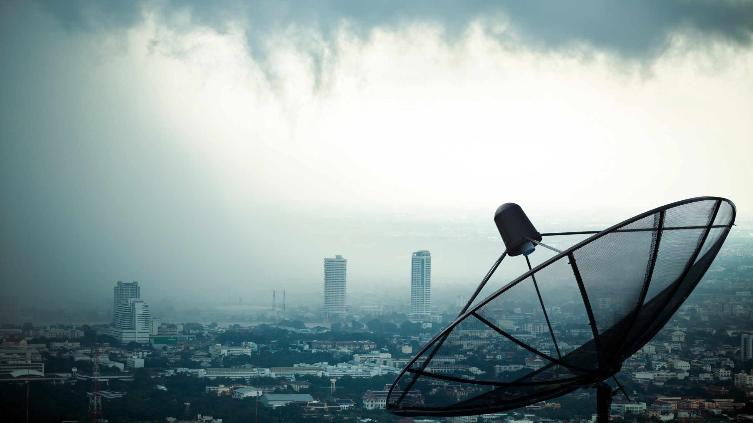 Satellite dish on top of a building in a large city with skyscrapers and other large buildings; the sky is dark and cloudy.