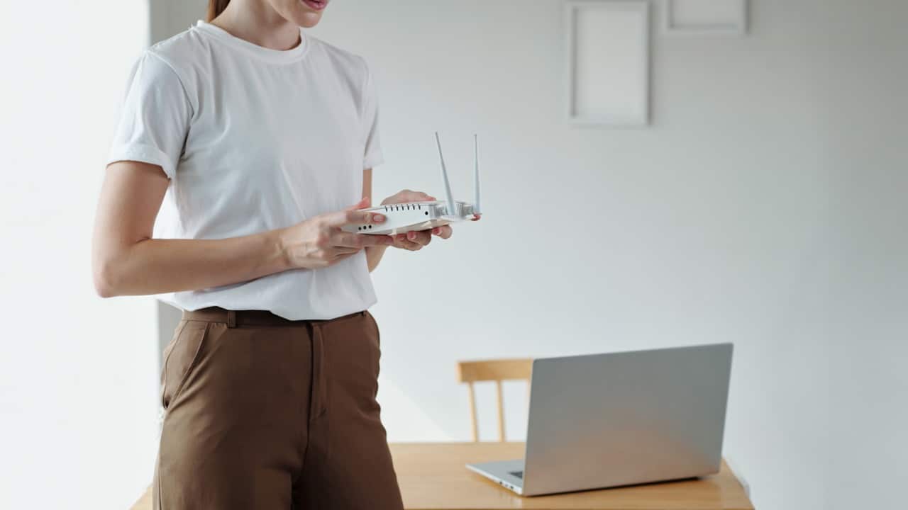 Woman holding a white Wi-Fi router near a laptop sitting on a wooden table
