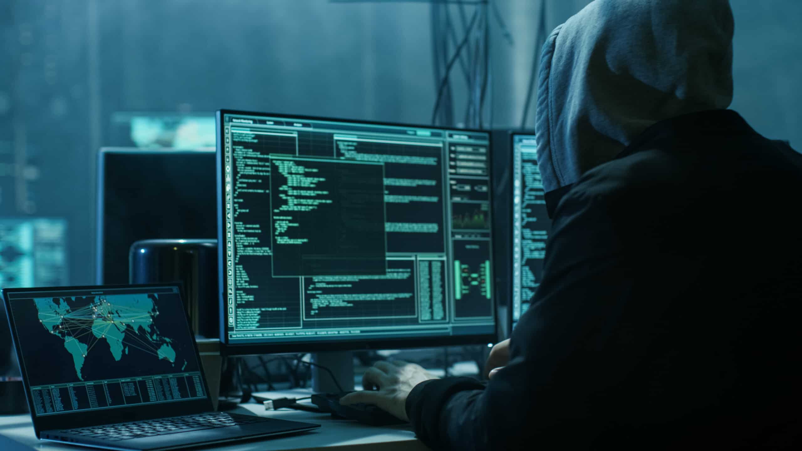 A hacker sitting at a desk wearing a black hoodie; he is typing on a monitor with several windows open with a laptop sitting next to it.