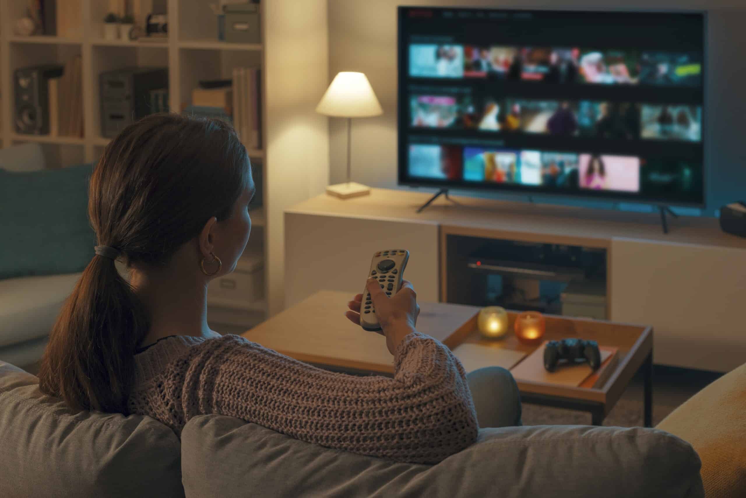 A woman holding a TV remote while sitting on the couch in front of a TV. The lighting is low, with candles and lamps lighting the space.