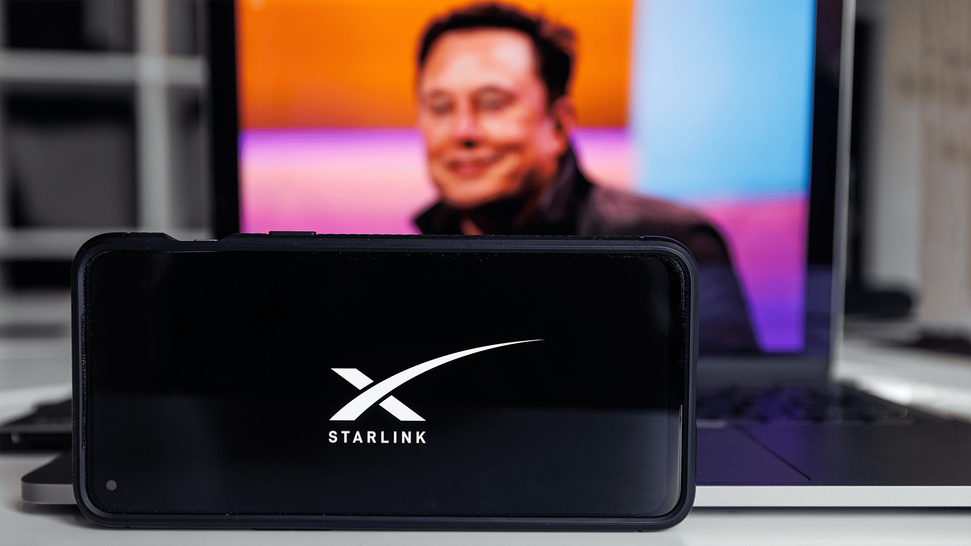 A smartphone with Starlink on it with Elon Musk’s picture on a laptop in the background.