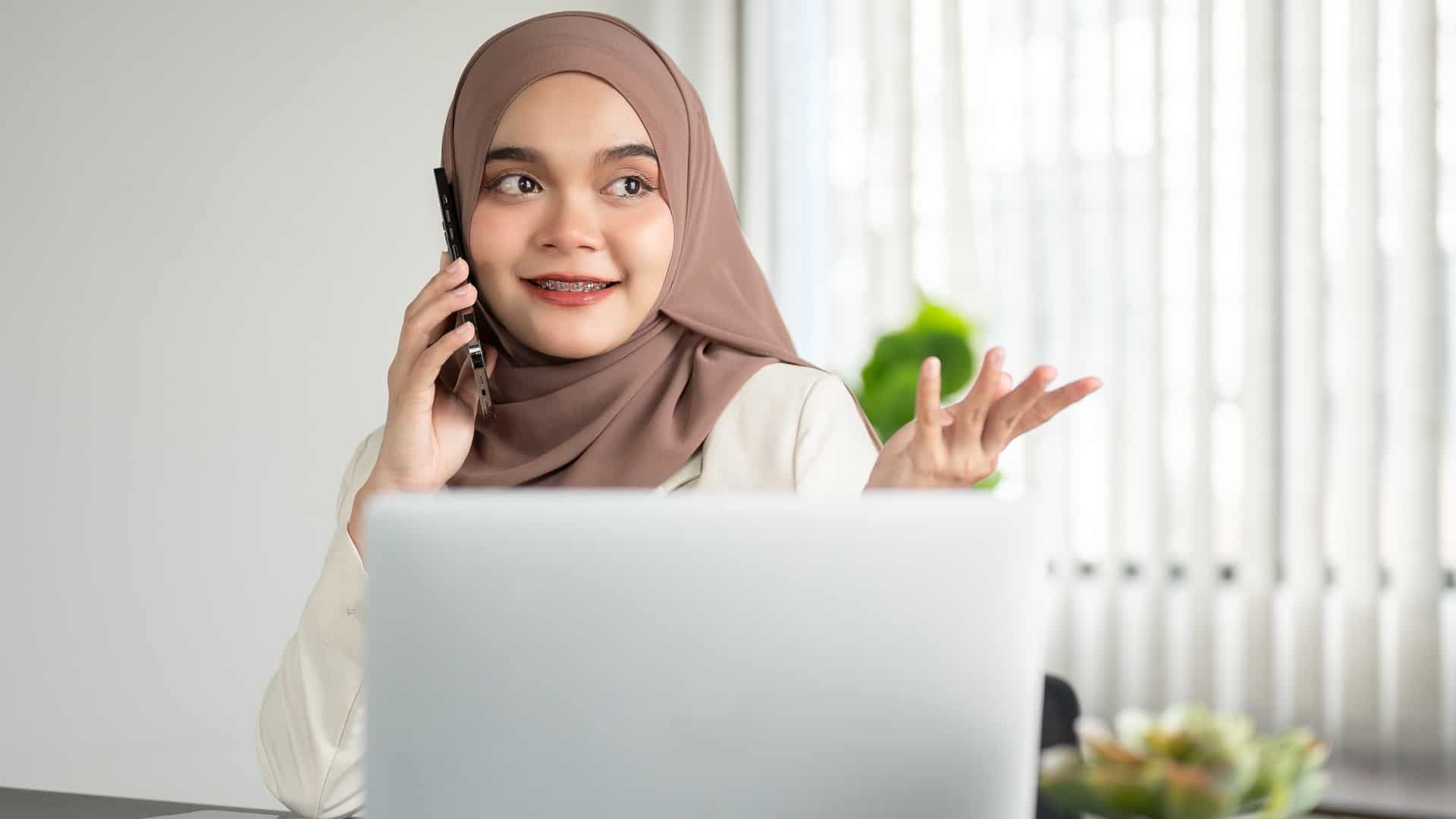 A woman talking on the phone while sitting in front of a laptop in this image from Shutterstock.