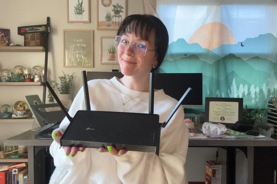 BroadbandNow Writer Hanna Shemke holding a black TP-Link Archer BE230 in front of a work desk.